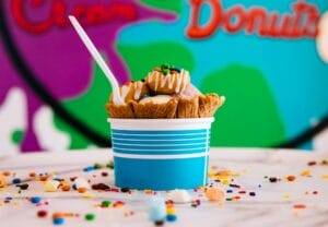 A scoop of ice cream in a blue and white container with a waffle cone piece, sprinkles, and a spoon on top. Multicolored sprinkles are scattered on the surface around it.