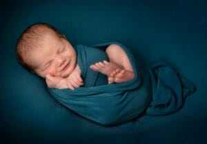 Newborn baby wrapped in teal fabric, sleeping peacefully against a matching teal background.