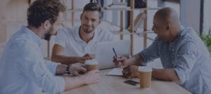Three men sit around a table with laptops and notepads, engaged in conversation. Two coffee cups are on the table.