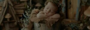A sleeping baby wearing a floral headband rests on a burlap-covered bucket. Surrounding foliage and soft lighting create a serene atmosphere.