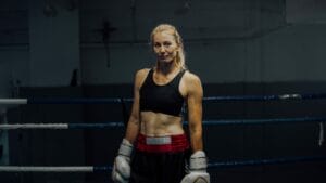 A woman in a sports bra and shorts stands confidently in a boxing ring, wearing boxing gloves.