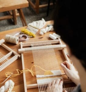 Person weaving with a wooden loom, using white and yellow yarn. Spools of thread and crafting materials are scattered on the wooden table.