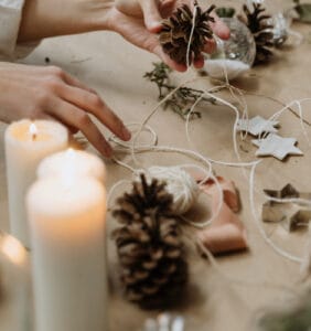 Hands arranging pinecones with twine on a table, surrounded by candles, star-shaped ornaments, and greenery.