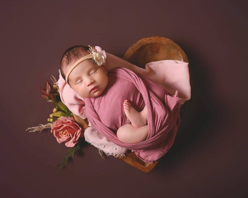 A sleeping baby wrapped in a pink cloth rests in a heart-shaped wooden bowl, surrounded by pink roses on a dark background.