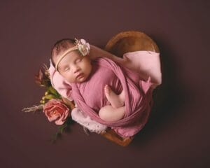 A sleeping baby wrapped in a pink cloth rests in a heart-shaped wooden bowl, surrounded by pink roses on a dark background.