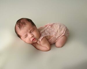 Newborn baby in pink lace outfit asleep on a soft surface against a neutral background.