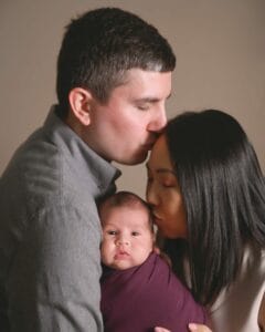 A man kisses a woman's forehead while she kisses a baby's cheek. The baby, wrapped in a purple blanket, looks at the camera.