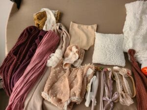 A collection of baby clothing and accessories, including lace outfits, fabric wraps, headbands, and a cushion, arranged on a beige surface.