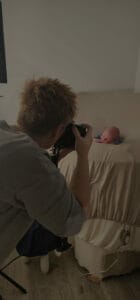 Person taking a photo of a newborn baby lying on a bed with beige sheets.