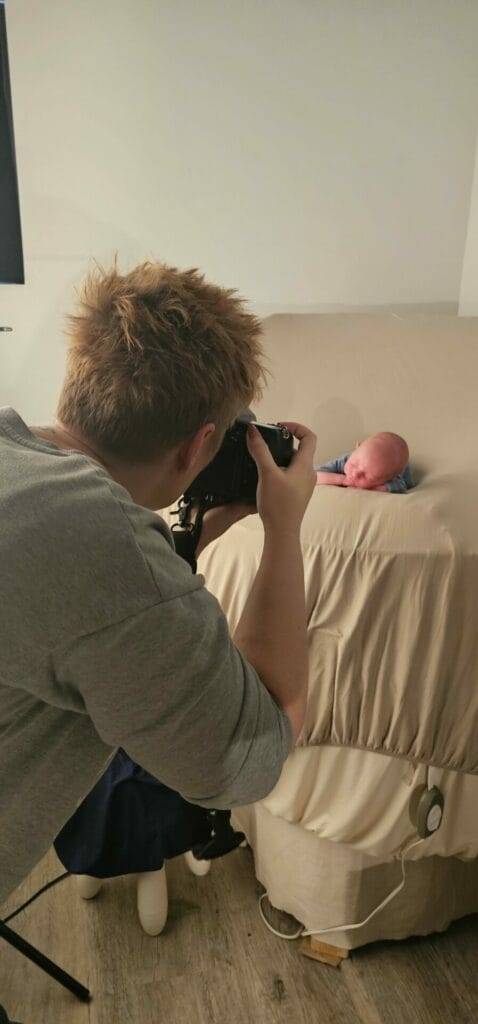 Person with short hair taking a photo of a baby lying on a beige blanket covering a table.