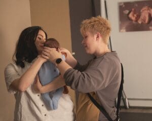 Two people are adjusting a baby wrapped in a blue blanket. One person holds the baby while the other adjusts the covering. A photograph of a baby is visible on the wall behind them.