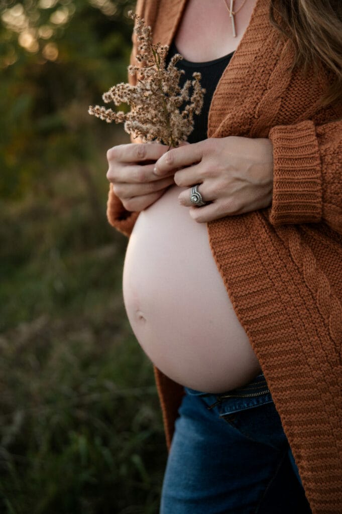 A pregnant person stands outdoors, gently holding dried flowers. They wear a brown knit cardigan and jeans, with a visible pregnant belly.