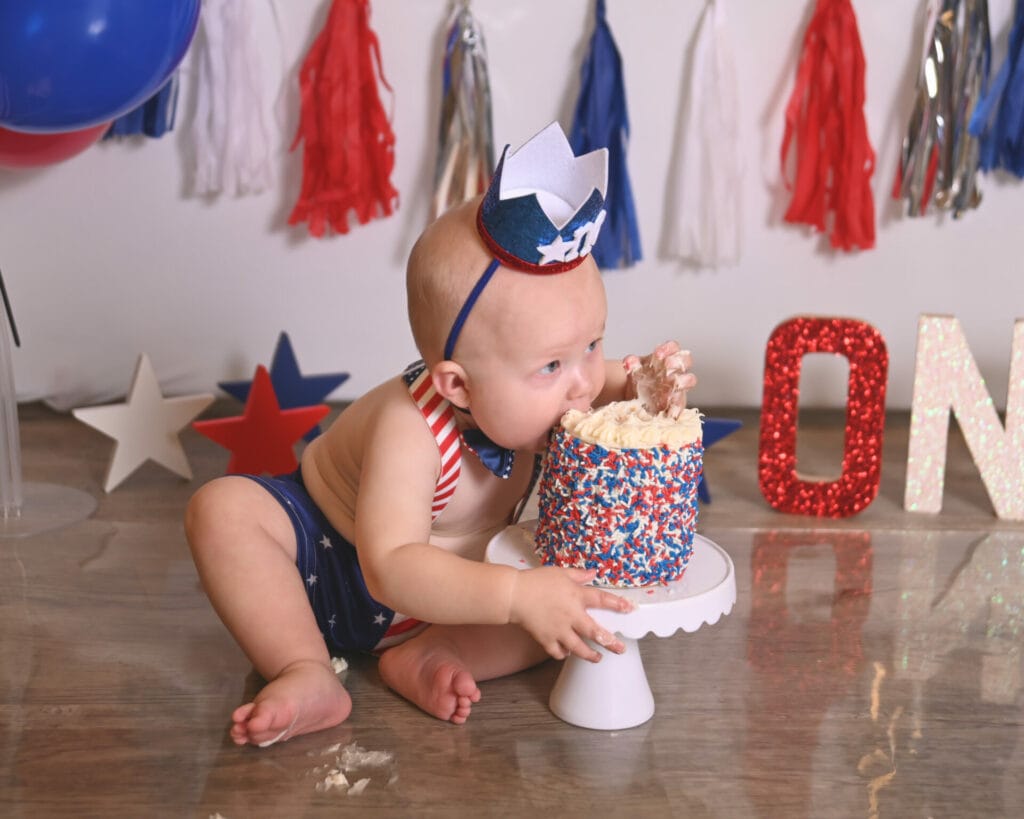 Baby in star-themed outfit and hat sits on wooden floor, eating a cake with sprinkles. Red, white, and blue decorations and a large "ONE" sign are in the background.