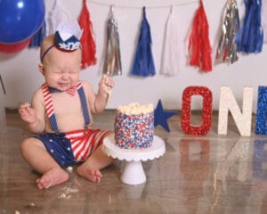 A baby in a red, white, and blue outfit sits on the floor, crying beside a small, decorated cake. The background features colorful tassels and the word "ONE" in large letters.