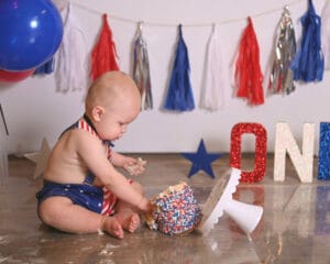 A baby in a patriotic outfit sits on the floor, playing with a cake covered in red, white, and blue sprinkles. A banner and decorations in matching colors are in the background.
