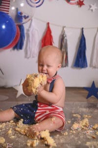 A baby in patriotic attire sits on the floor with cake smeared around, holding a large piece. Balloons and festive decorations are in the background.