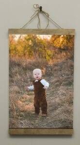 A baby in brown overalls and a white shirt stands on a dirt path in a grassy field with autumn foliage in the background, framed and hanging on a wall with twine.
