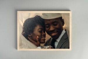A smiling couple poses closely, wrapped in a light fabric. The woman has short curly hair, and the man is wearing a suit. The image is framed with a light wooden border.