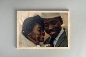 A joyful couple embraces under a white cloth, framed in a wooden picture frame placed on a gray surface.