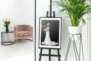 Framed photo of a bride on an easel next to a potted plant, with a pink chair and flowers on a table in the background.
