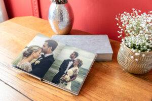 An open photo album on a wooden table shows a couple in wedding attire. A vase with white flowers and a silver decorative item are in the background against a red wall.