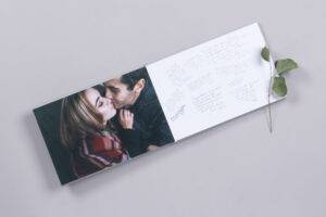 Open photo album showing a couple kissing on the left page and handwritten messages on the right, with a sprig of leaves resting on the open pages.