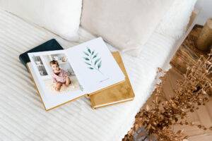 Open photo book displaying a baby and leaf illustration, placed on a bed with cushions and a nightstand with a candle and dried flowers.
