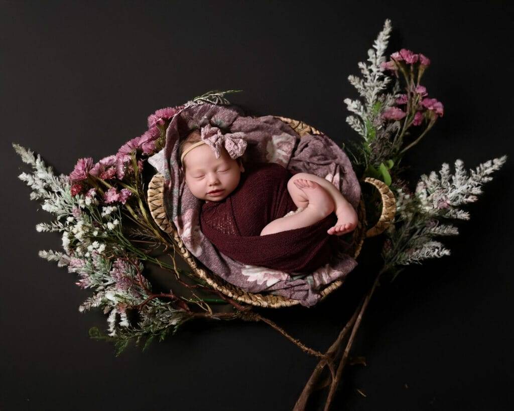 Sleeping baby in a maroon wrap with a bow on head, lying in a basket surrounded by purple and white flowers on a dark background.