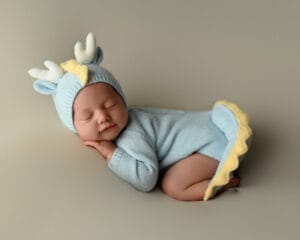 Baby sleeping peacefully in a blue and yellow outfit with a matching hat featuring horns. The baby is lying on their stomach with hands under the chin. Soft, neutral background.