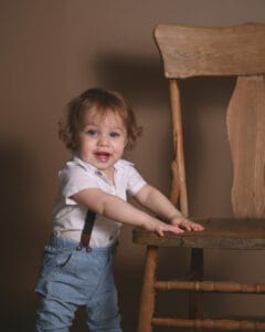A toddler in a white shirt and suspenders stands beside a wooden chair, smiling at the camera against a plain background.