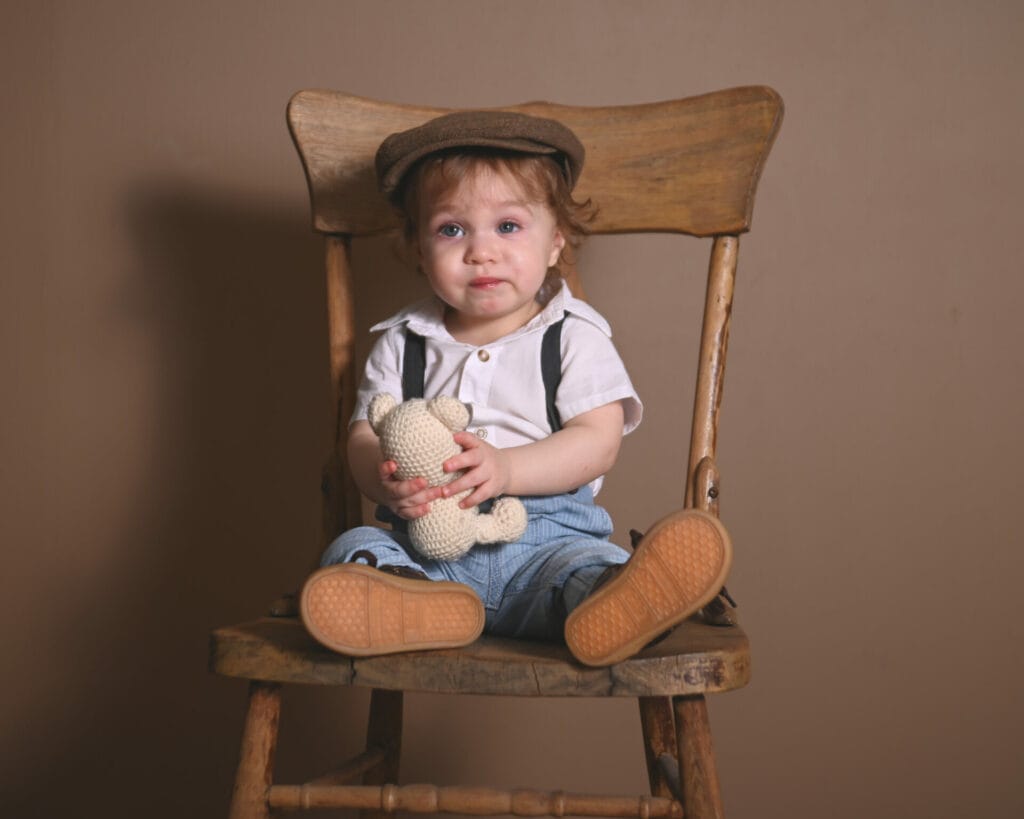 Young child in vintage attire sits on a wooden chair holding a knitted teddy bear against a brown backdrop.