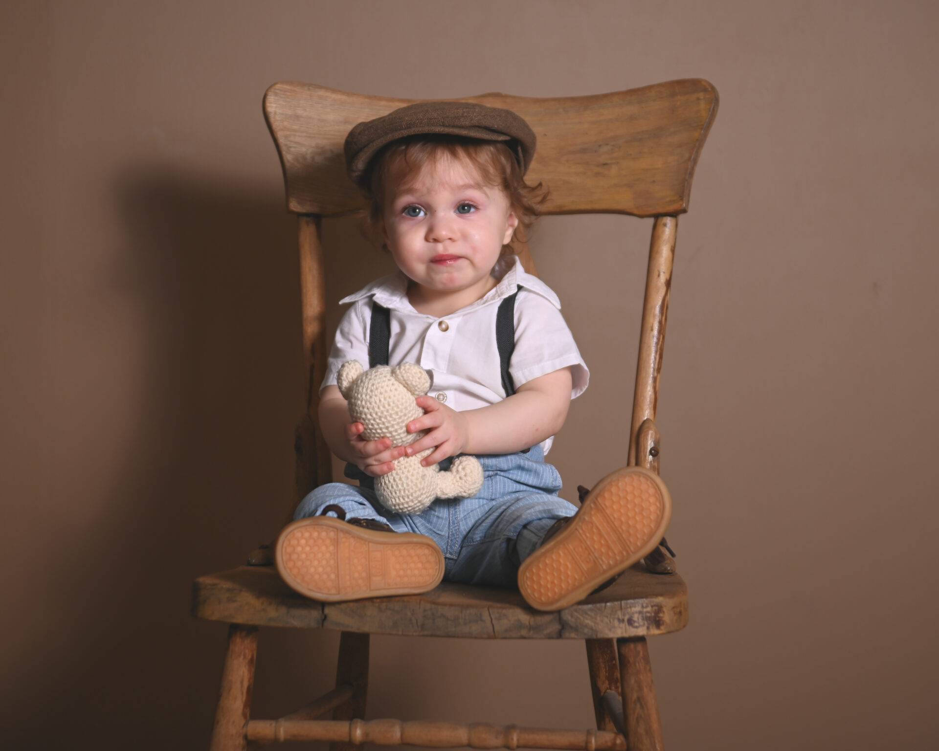 Young child in vintage attire sits on a wooden chair holding a knitted teddy bear against a brown backdrop.