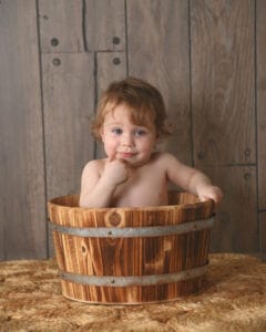 A toddler sits in a wooden bucket, resting one hand on the edge and the other near their face, against a wooden backdrop.