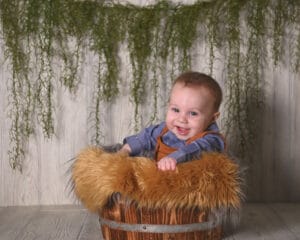 A baby smiling in a wooden bucket with furry padding, set against a wooden backdrop and hanging green foliage.