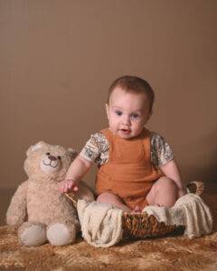 A baby in an orange outfit sits in a basket holding a teddy bear on a textured surface against a plain brown background.