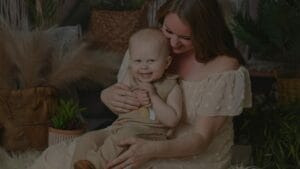 Woman in a light dress holds a smiling baby in a beige outfit, seated among plants and baskets.