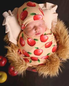 A baby dressed in apple-patterned clothing sleeps in a basket lined with faux fur, surrounded by red and yellow apples.