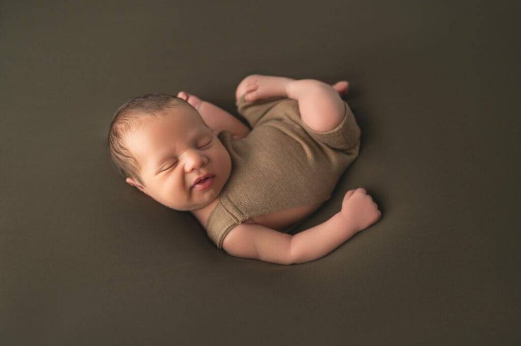 Newborn baby sleeping on their side, dressed in a brown sleeveless outfit, against a plain dark background.