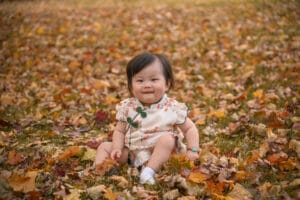 A baby in a floral outfit sits on a lawn covered with autumn leaves, surrounded by scattered red, yellow, and brown foliage.