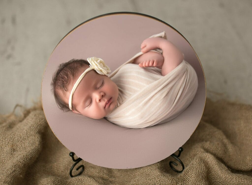 A newborn baby sleeps peacefully, wrapped in a cream swaddle with a matching headband with a flower, displayed in a circular frame on a stand.