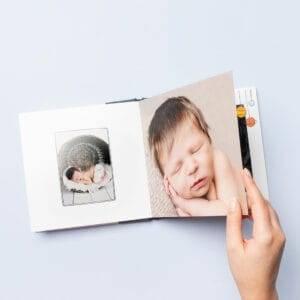A hand holds open a photo album displaying images of a sleeping newborn baby on the left and right pages against a light blue background.