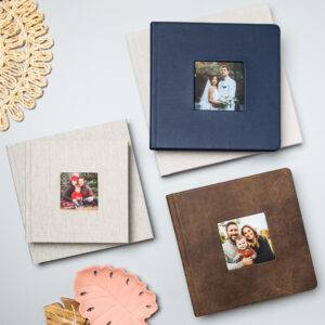 Four photo albums with pictures on the covers are arranged on a gray surface, with a woven mat and decorative leaves nearby.