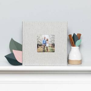 A photo album with a linen cover featuring a couple's picture is displayed on a shelf next to a vase with leaves and cinnamon sticks.