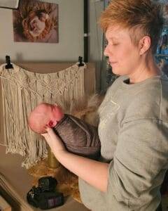 Person holding a wrapped newborn in a photography studio with a camera nearby and a macramé backdrop.
