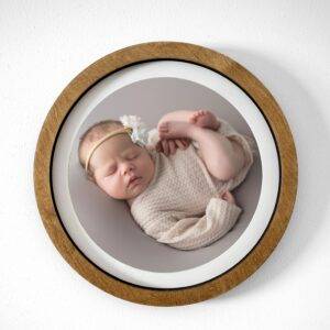 A newborn baby wearing a headband and beige outfit is sleeping, curled up in a circular frame against a white background.