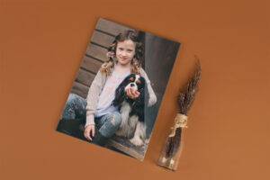 A framed photo of a young girl seated with a black and white dog on a brown wooden background, next to a small clear vase with dried grass, placed on an orange surface.