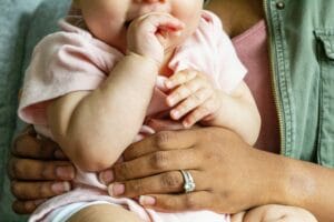 An adult holding a baby wearing a pink outfit. The adult's hand, adorned with a ring, supports the baby who is sucking on their fingers.