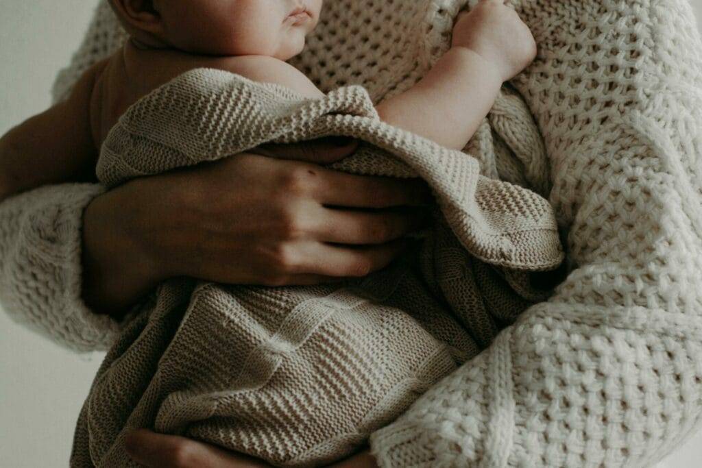 An adult wearing a knitted sweater holds a baby wrapped in a knitted blanket.