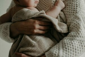 An adult wearing a knitted sweater holds a baby wrapped in a knitted blanket.