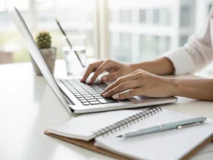 Person typing on a laptop at a bright desk, with a notebook, pen, and small plant nearby.